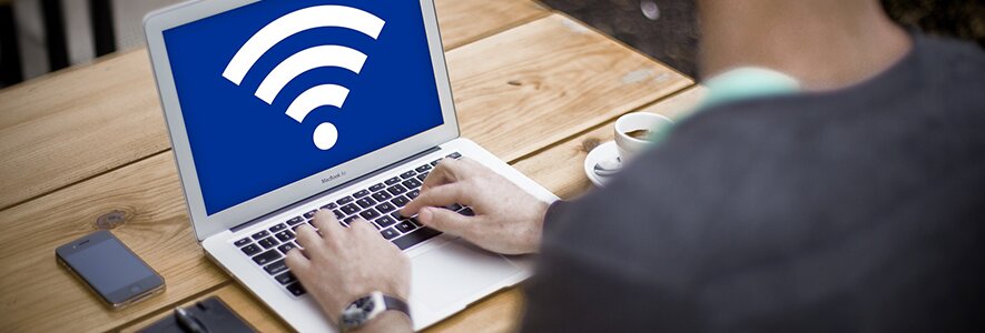 Eine Person sitzt an einem Holztisch mit Laptop, auf dessen Bildschirm ein großes weißes WLAN-Symbol auf blauem Hintergrund zu sehen ist.