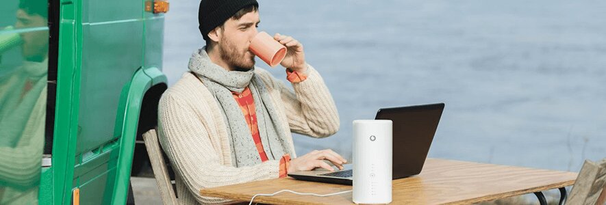  Ein Mann mit Mütze und Schal sitzt an einem Holztisch im Freien, trinkt aus einer Tasse und nutzt einen Laptop, während ein LTE-Router daneben steht.