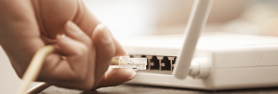 Eine Hand verbindet ein gelbes Ethernet-Kabel mit einem weißen WLAN-Router, der eine Antenne an der Seite hat.