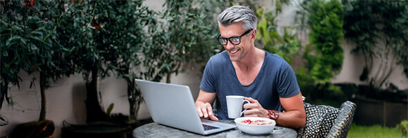 Ein grauhaariger Mann mit Brille sitzt draußen an einem Tisch, arbeitet an einem Laptop und hat eine Tasse Kaffee und eine Schüssel Müsli neben sich.