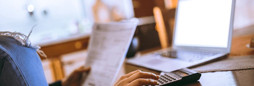 Eine Frau hält eine Rechnung in der einen Hand und tippt mit der anderen auf einen Taschenrechner. Im Hintergrund ein Laptop mit heller Bildschirmbeleuchtung.