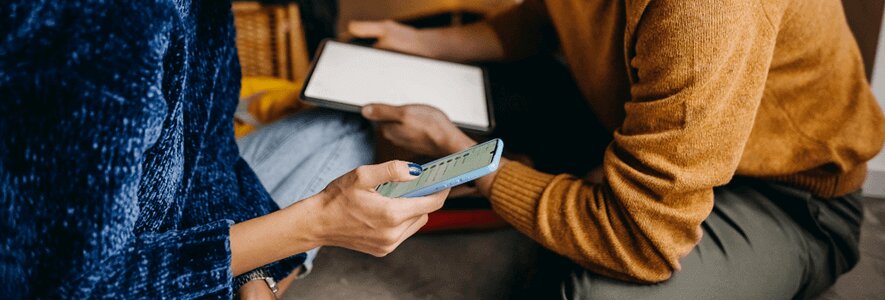  Eine Frau in blauem Pullover hält ein Smartphone in der Hand, während ein Mann in braunem Pullover daneben ein Tablet benutzt.