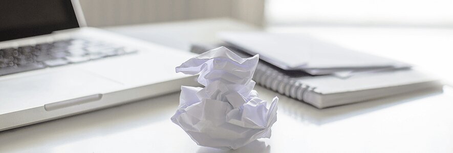 Ein zerknülltes Stück Papier liegt auf einem weißen Schreibtisch neben einem Laptop und einem Notizblock.