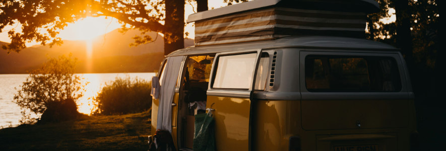 Ein gelbes Retro-Wohnmobil mit aufgeklapptem Dach steht am Ufer eines Sees. Die Sonne geht im Hintergrund unter und taucht die Szene in warmes Licht.