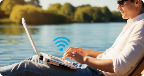 Mann mit Sonnenbrille arbeitet entspannt am Laptop am Seeufer mit WLAN-Symbol.