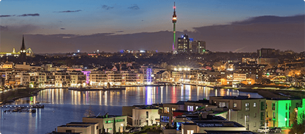 Nachtaufnahme des Phoenix-Sees in Dortmund mit beleuchteten Häusern und dem Florian-Turm im Hintergrund.